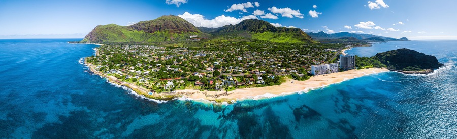 Vista aérea de un pueblo costero con playa, aguas azules cristalinas y montañas verdes exuberantes al fondo.
