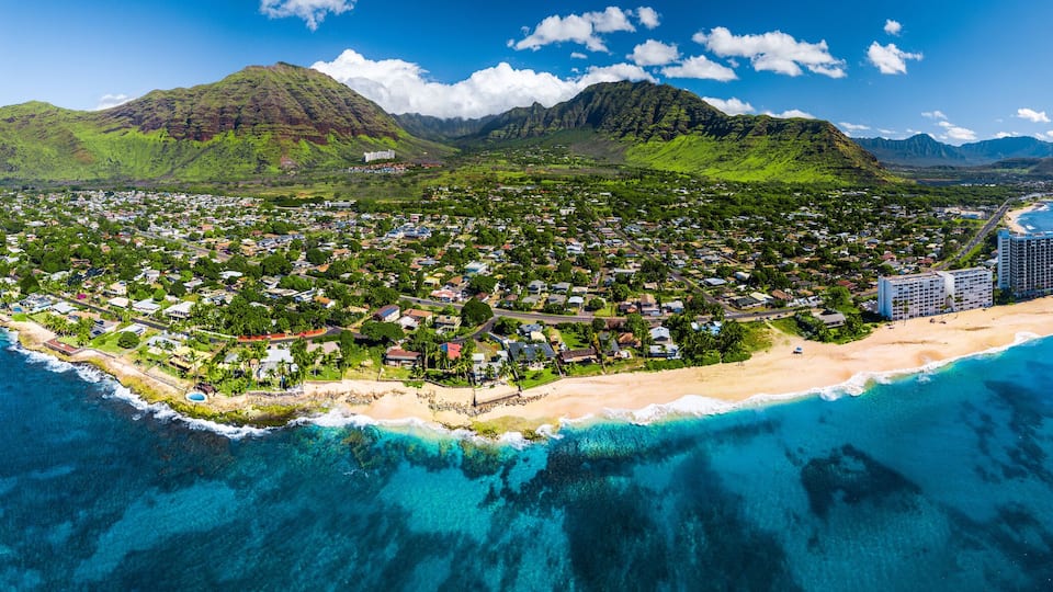 An aerial view of a coastal town with a beach, clear blue waters, and lush green mountains in the background.