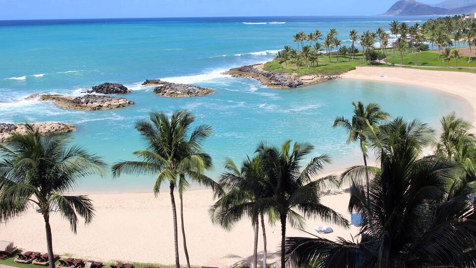 A tropical beach with clear turquoise waters, palm trees, and a distant mountain range.