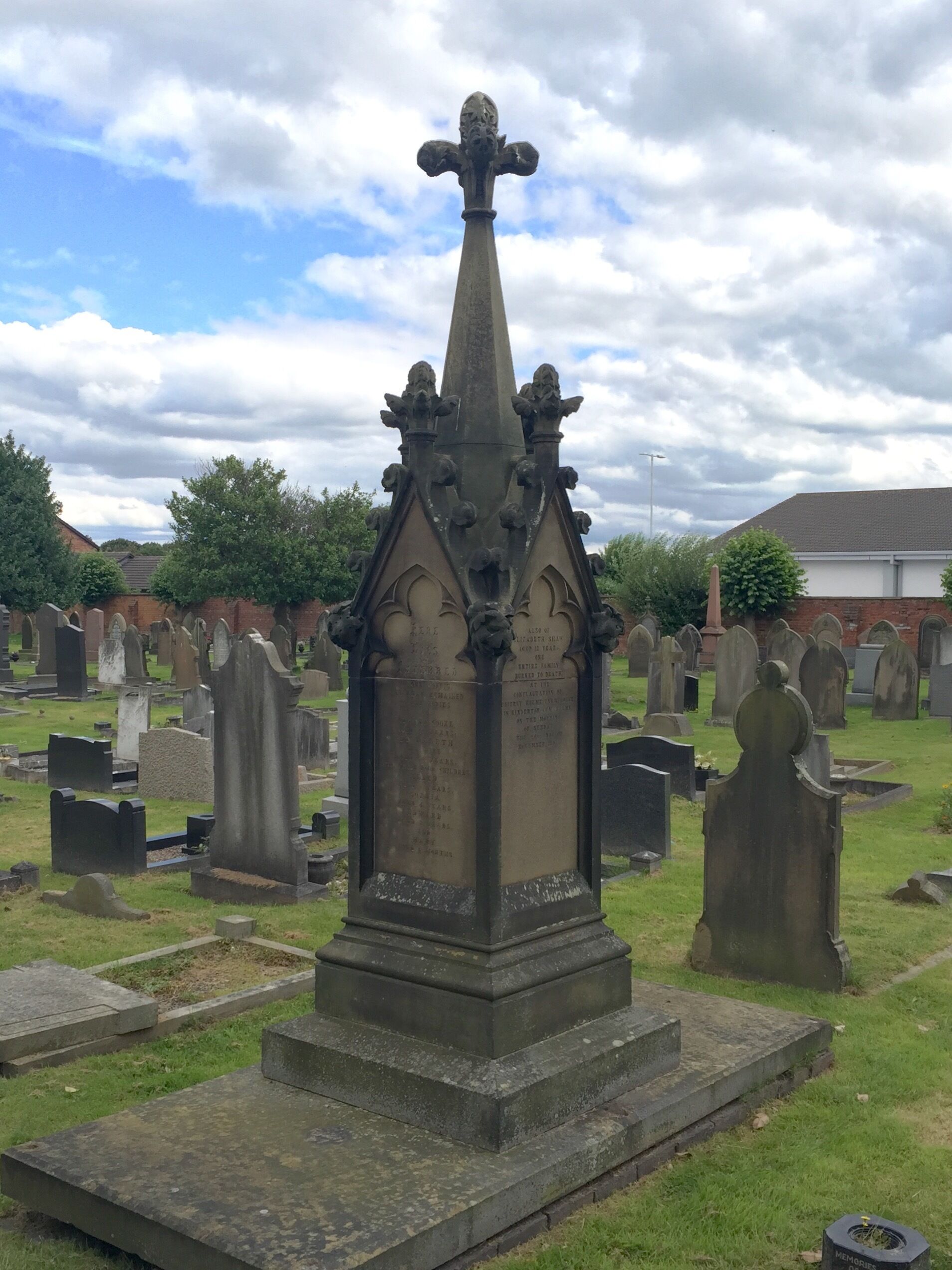 A stone monument consisting of a square base with inscribed panels, surmounted by a spire. The monument was paid for by public subscription, and commemorates seven members of a family killed in a house fire.