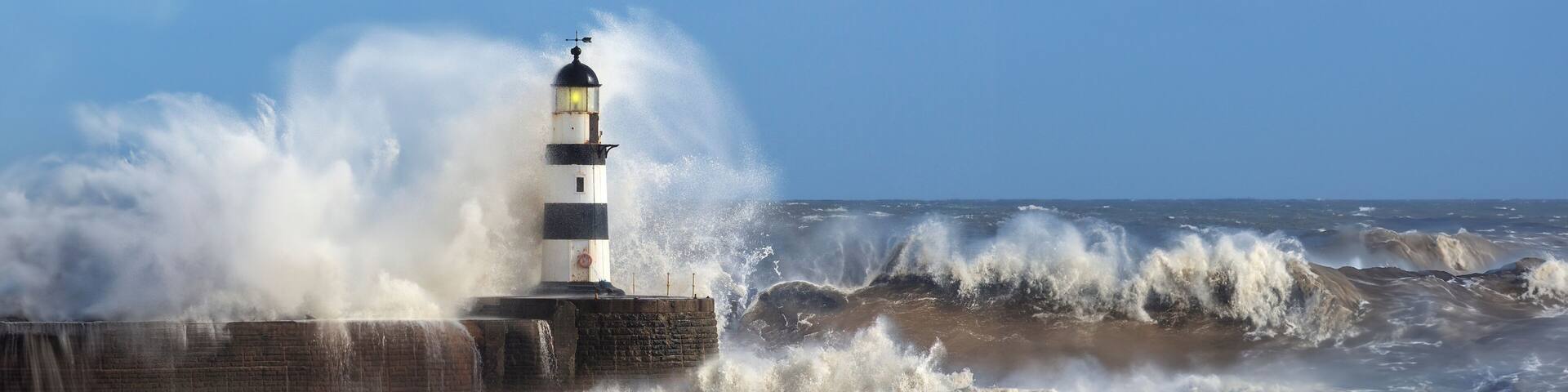 A lighthouse by a turbulent sea with waves crashing against a concrete barrier.