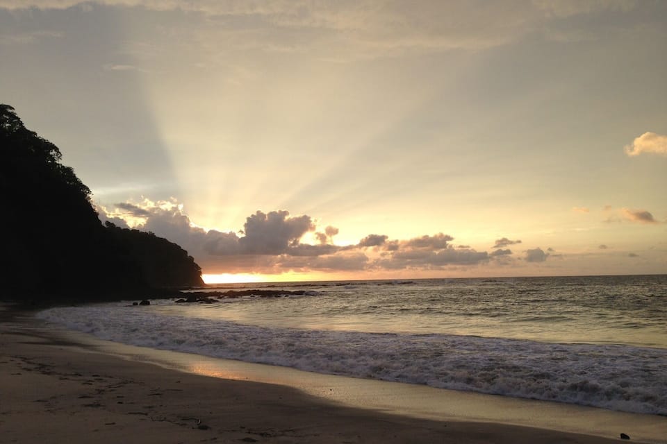 Una playa al atardecer con olas rompiendo sobre la orilla y un acantilado al fondo.