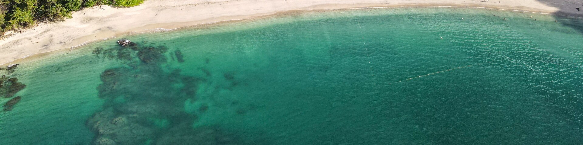 Vista aérea de uma praia com águas turquesa cristalinas, areal arenoso e área florestal densa.