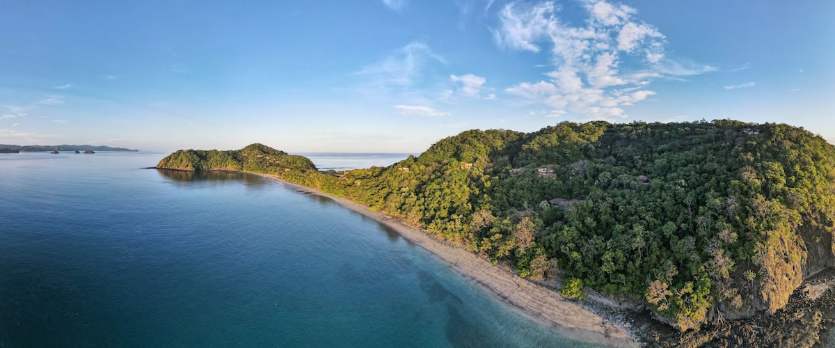 A coastal landscape with a beach, forested hills, and a clear sky.