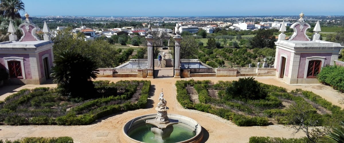 A landscaped garden with a central fountain, surrounded by manicured bushes and trees, leading to a gate with a view of a distant town.