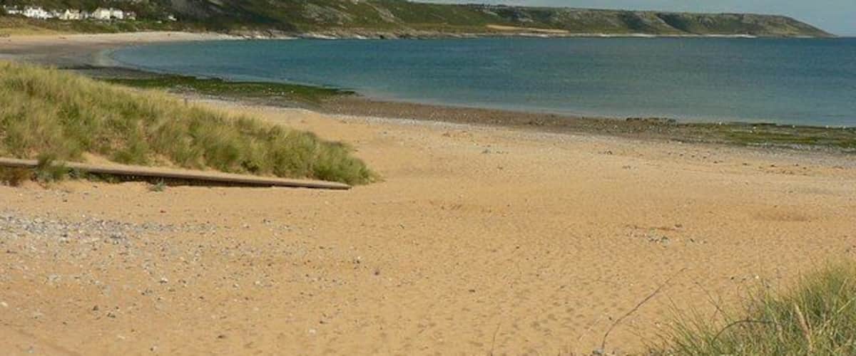 A sandy beach with a calm sea, grassy dunes, and a distant hill with houses.