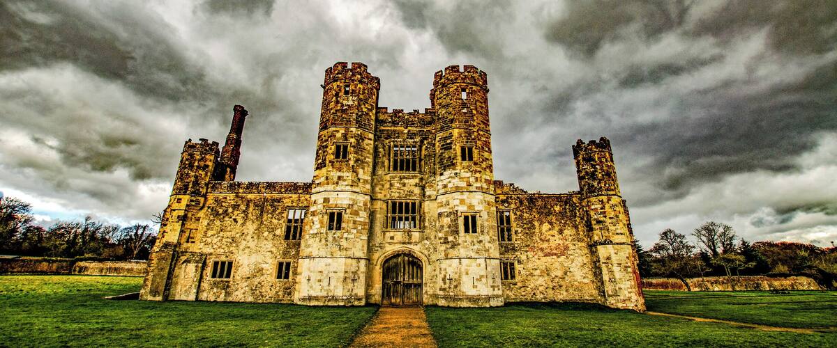 A historic castle with two towers, surrounded by a grassy field and under a cloudy sky.