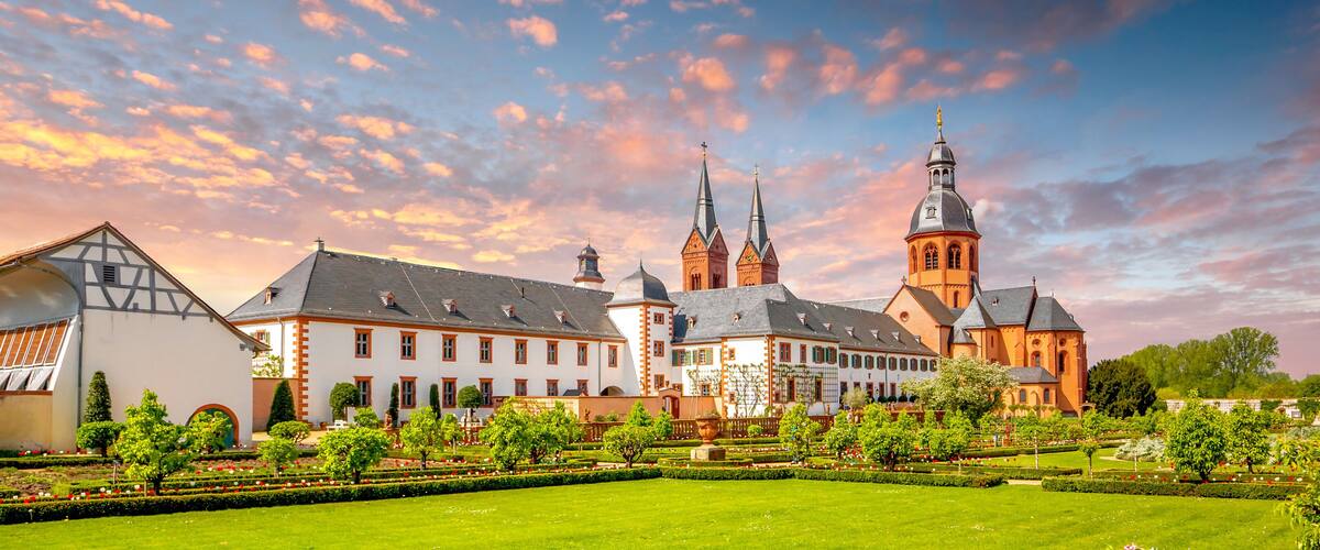 A historic building complex with a large tower, surrounded by well-maintained gardens and a clear sky.