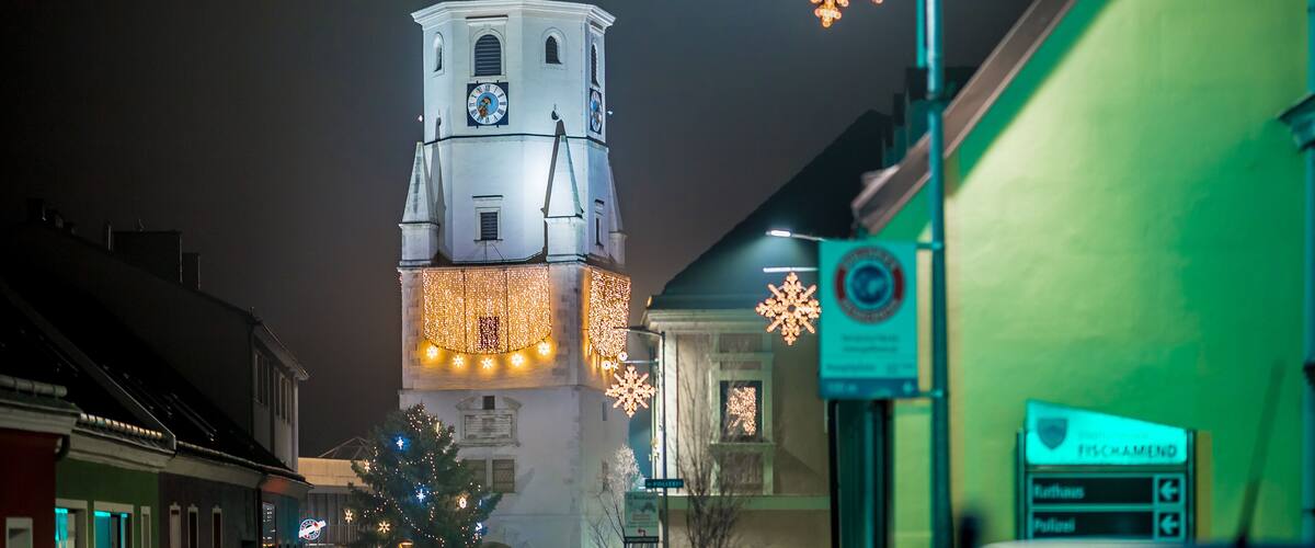 A night view of a town with a prominent clock tower adorned with lights, a street sign, and a green building.