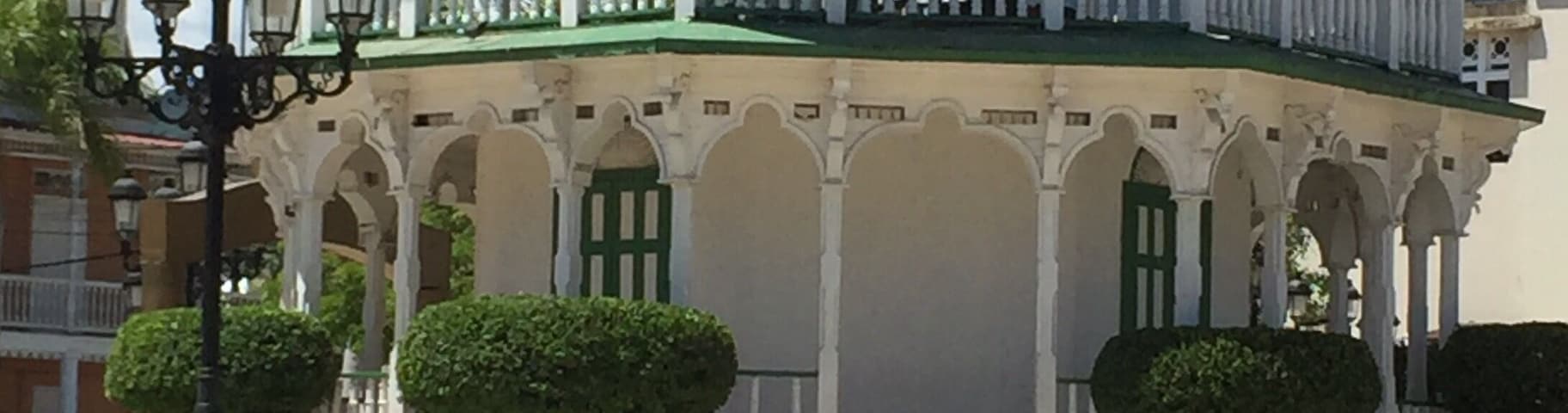 Ein weißes Gazebo mit grünem Zierstreifen und mehreren Bögen, umgeben von einem Zaun und Bänken.