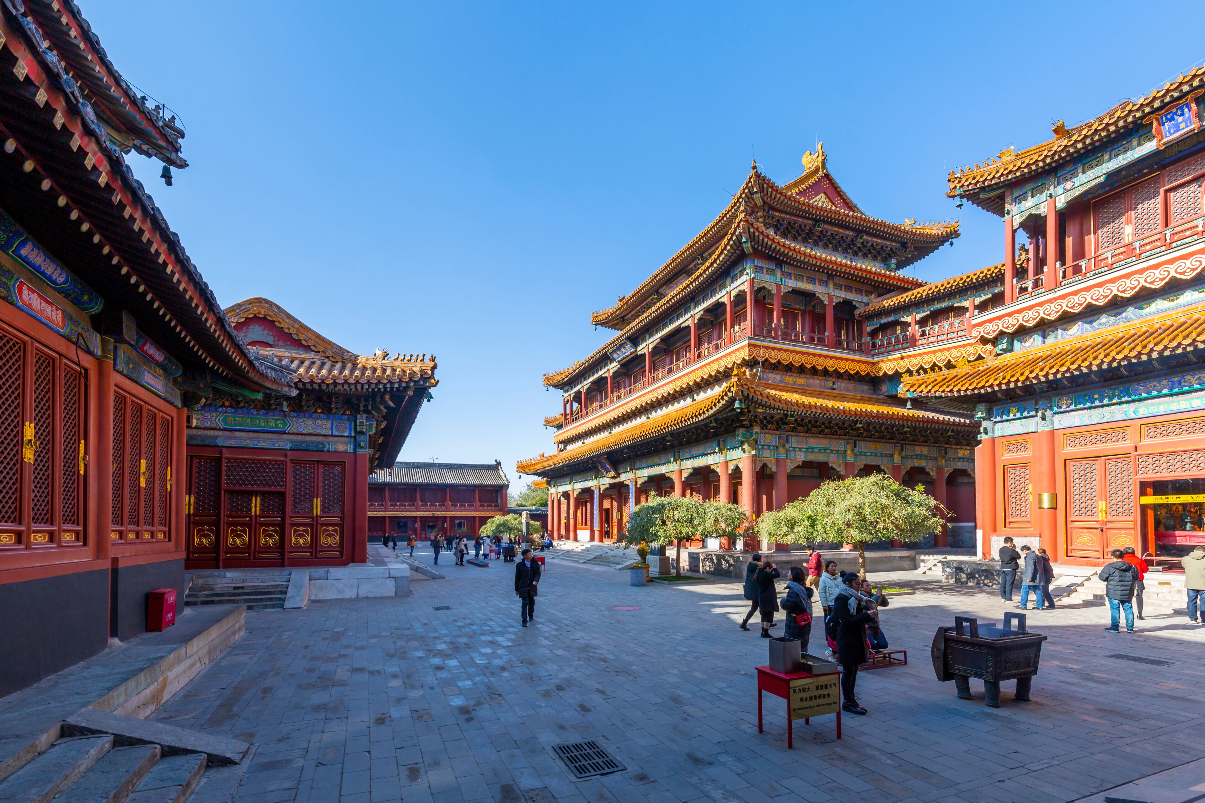 A traditional Chinese courtyard with ornate buildings, a clear blue sky, and people walking around.