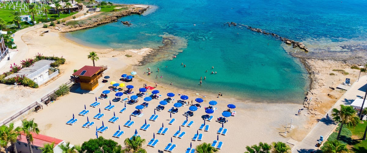 A coastal beach with clear turquoise waters, a rocky breakwater, and numerous sun loungers and umbrellas.