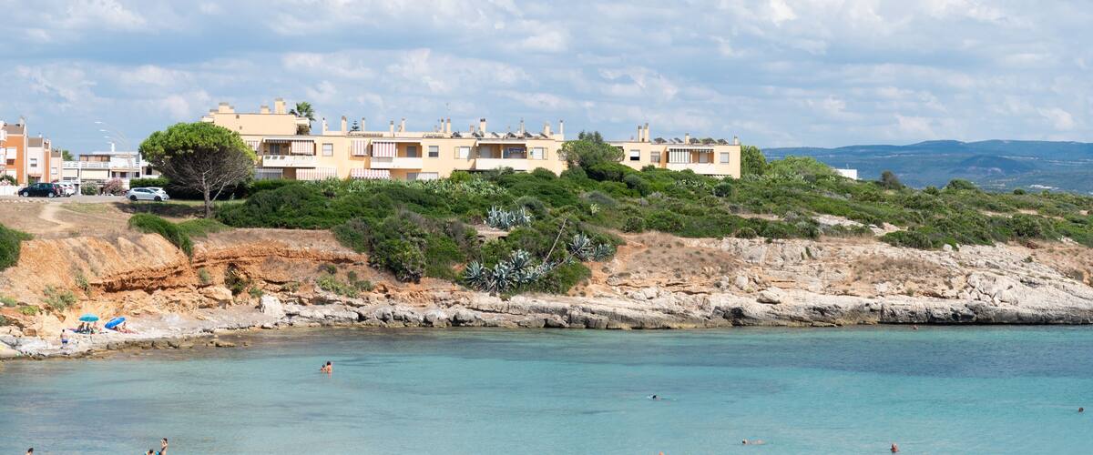 A coastal area with clear turquoise waters, rocky shoreline, and a row of beige buildings with red and white striped awnings.