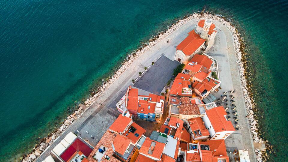 An aerial view of a coastal town with red-roofed buildings, a clear blue sea, and a rocky shoreline.