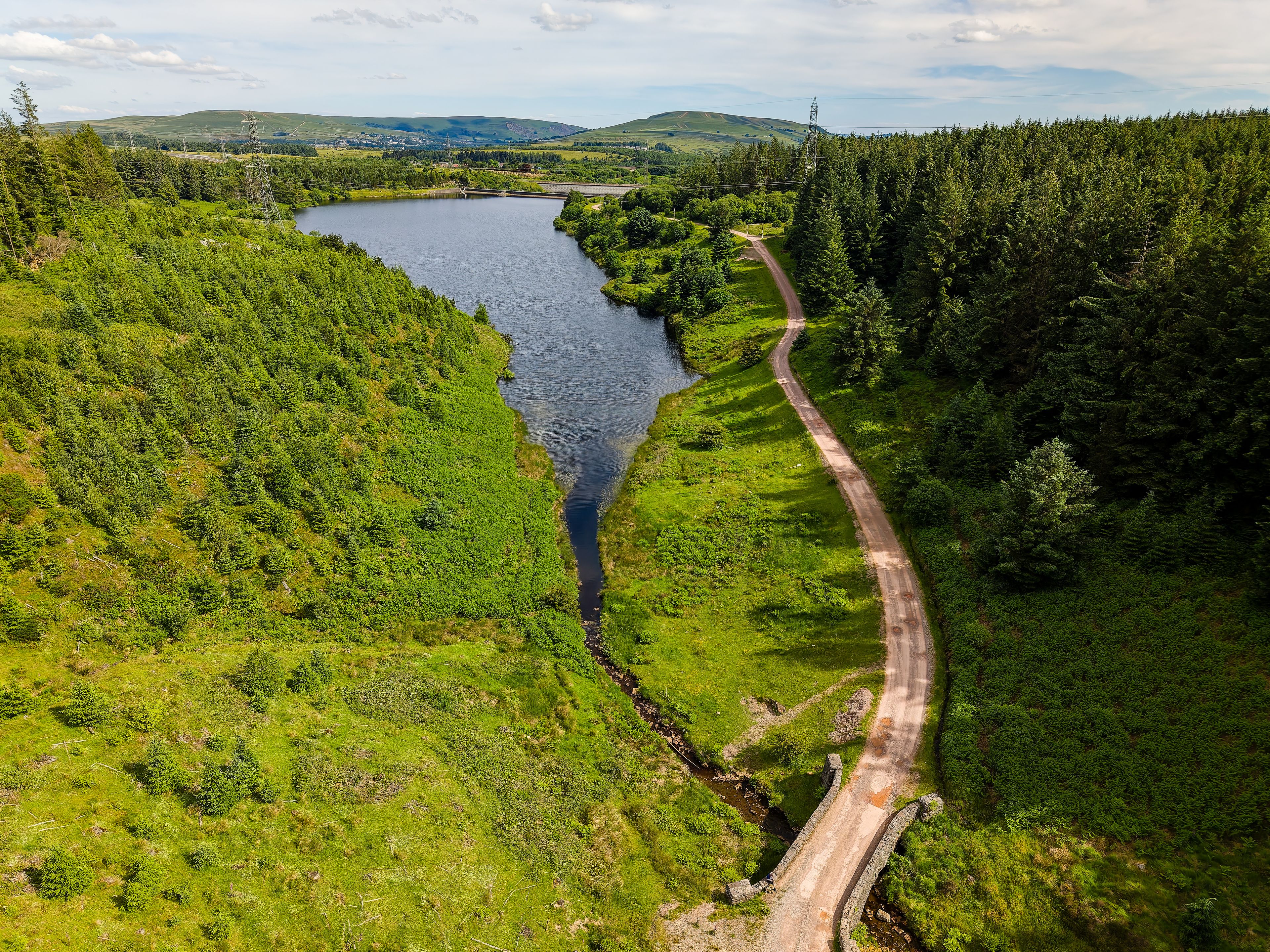 A winding road through a lush green landscape with a river and hills in the background.