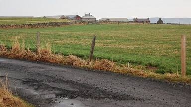 A rural road with a stone wall and wooden fence, leading to a field with houses in the background.