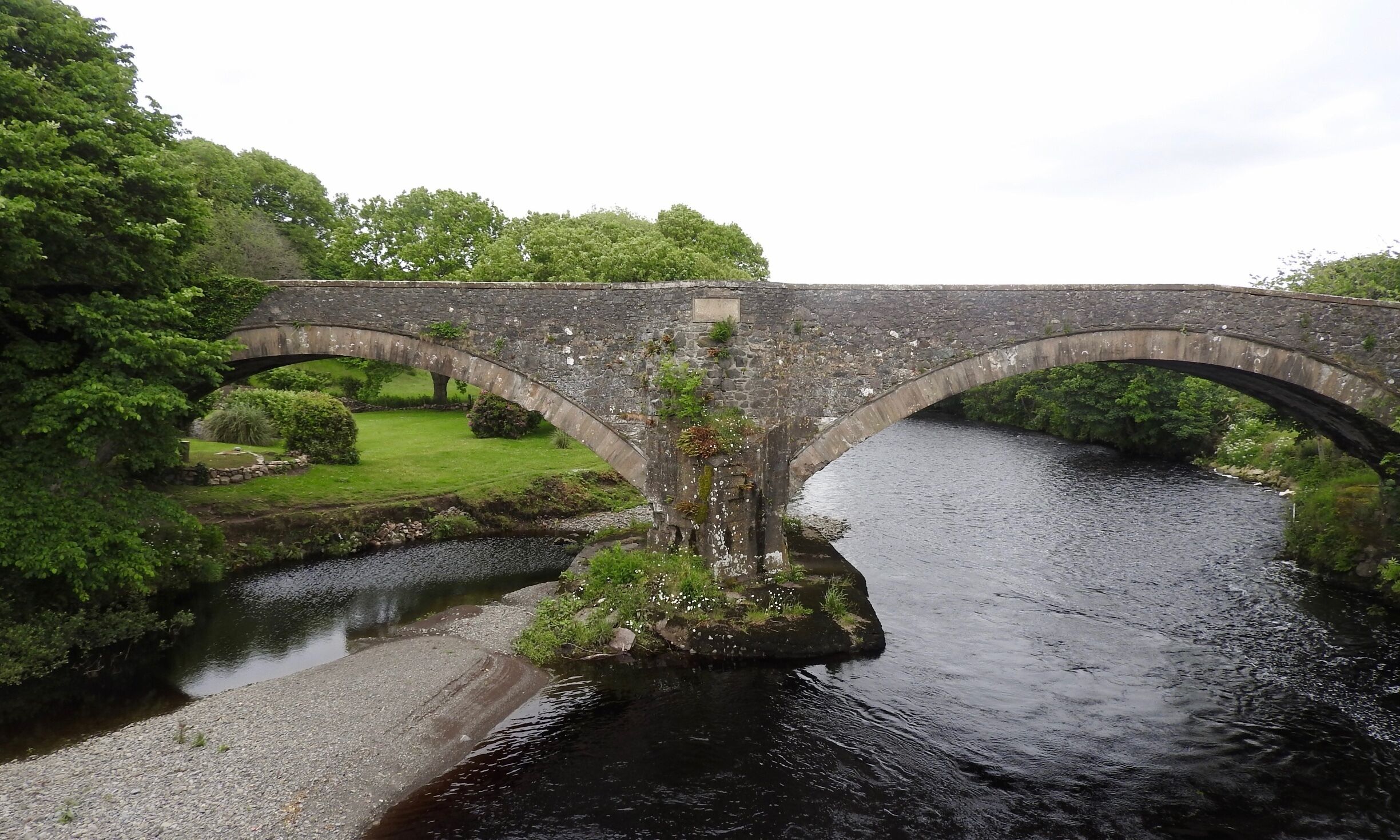 A stone bridge over a river with lush greenery and a well-maintained lawn.