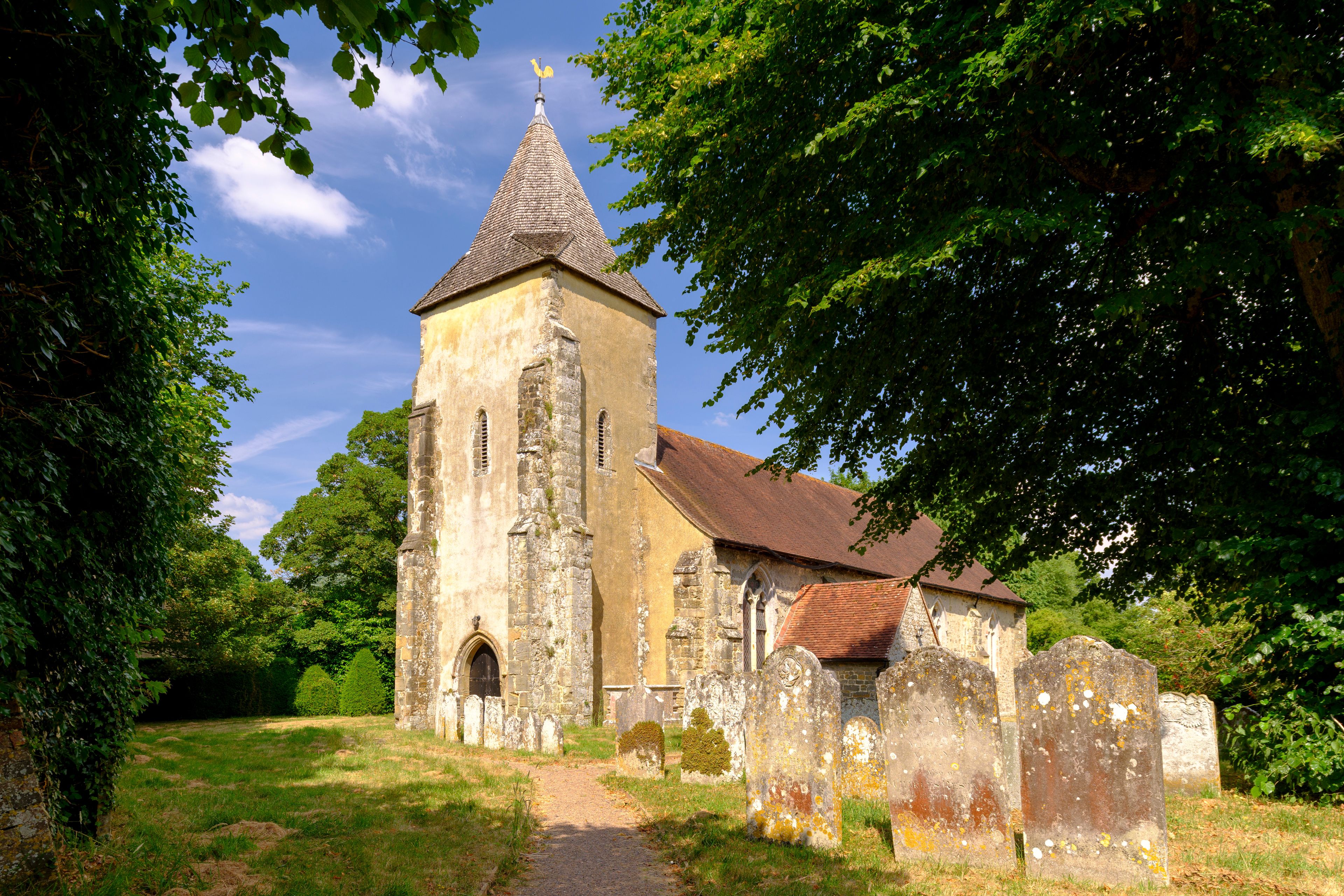 An old church with a pointed steeple, surrounded by trees and a grassy area with gravestones.
