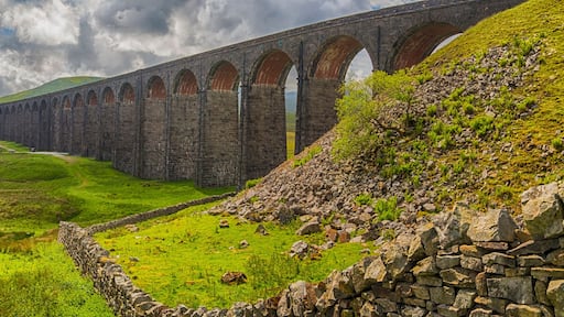 A viaduct over a valley with a winding road, green fields, and a cloudy sky.