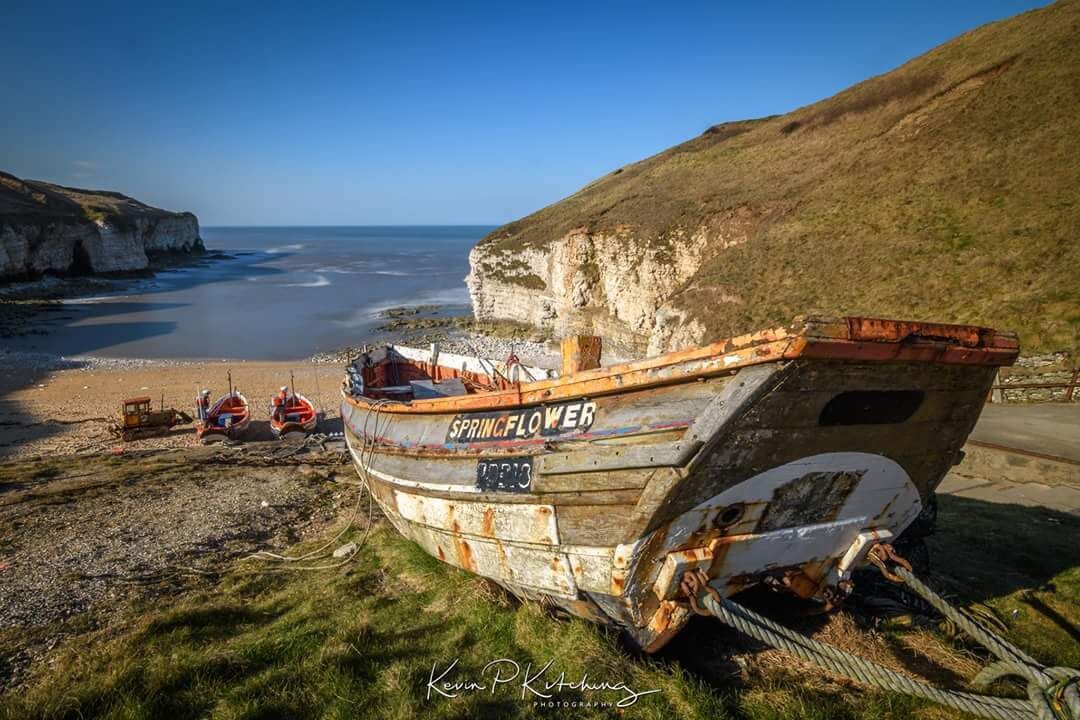 A weathered boat named SPRING FLOWER on a grassy beach with cliffs in the background.