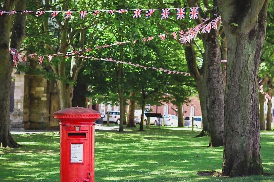 A red post box in a park with trees and a string of British flags.