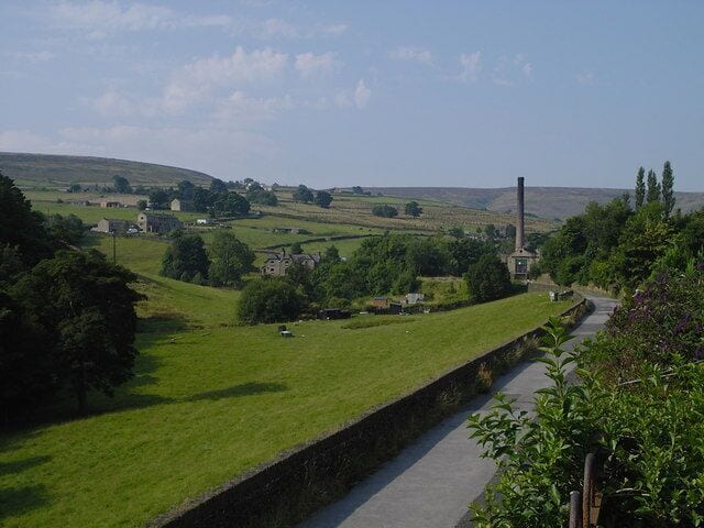 A rural landscape with a grassy field, a stone wall, a road, and a tall chimney.