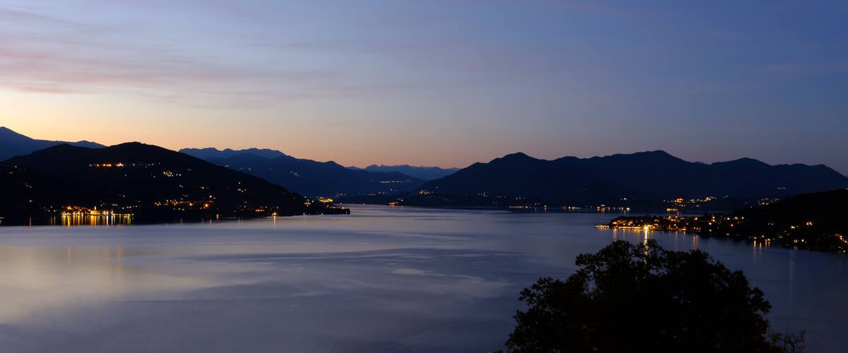 A calm lake at dusk with mountains in the background and city lights on the hills.
