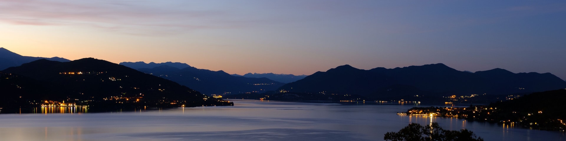 A calm lake at dusk with mountains in the background and city lights on the hills.