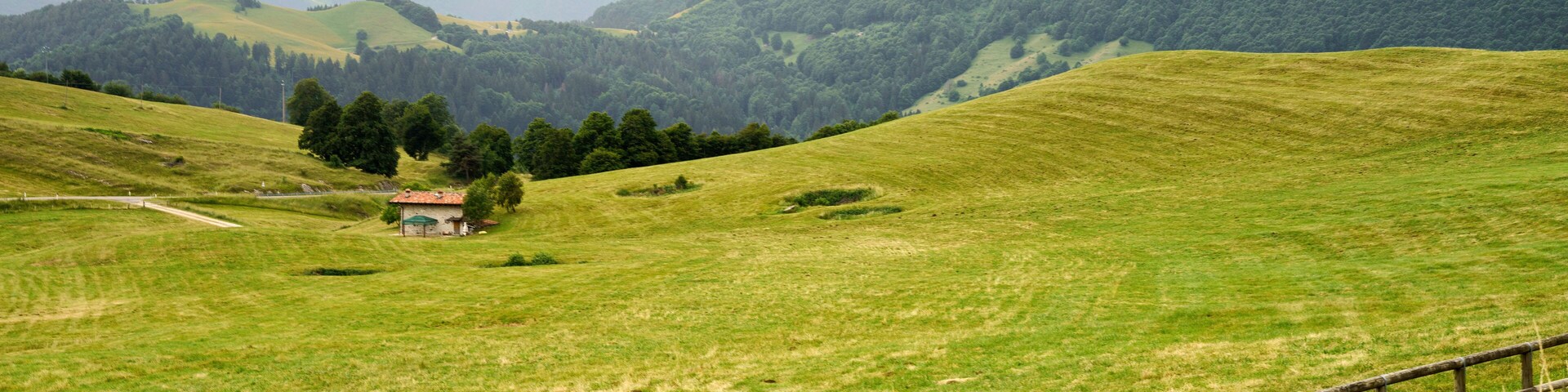 Un campo verde con una valla de madera, un edificio pequeño y montañas al fondo.