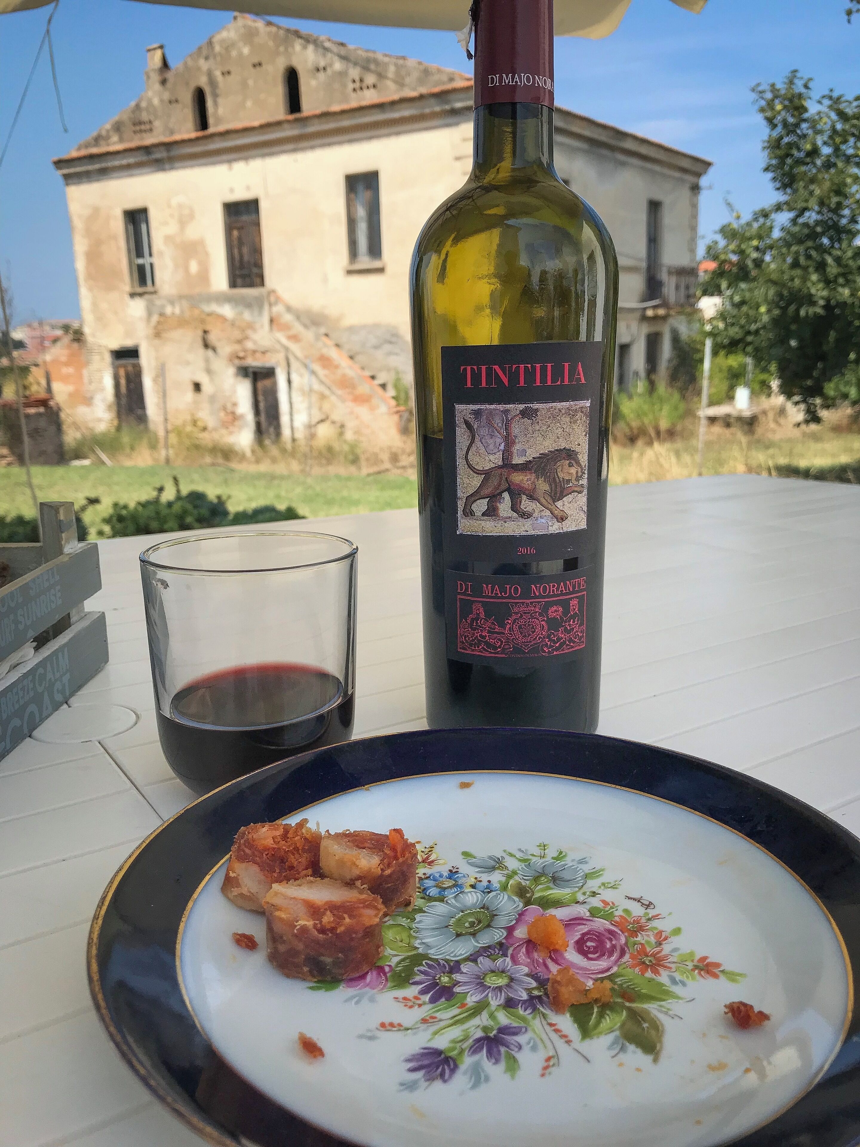 Eine Flasche Tintilia-Wein, ein Weinglas und ein Teller mit Speisen auf einem Tisch. Im Hintergrund ist ein Gebäude zu sehen.