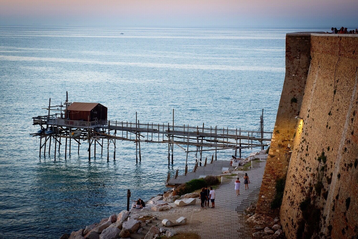 A pier extending into calm waters, a wooden structure on the pier, and a stone wall on the right.