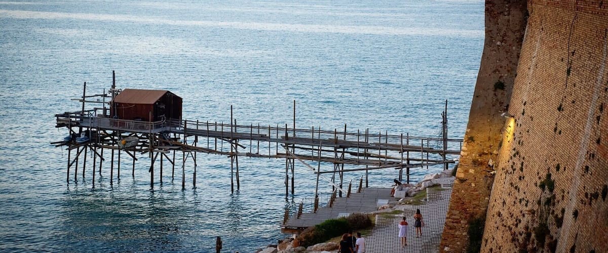 A pier extending into calm waters, a wooden structure on the pier, and a stone wall on the right.