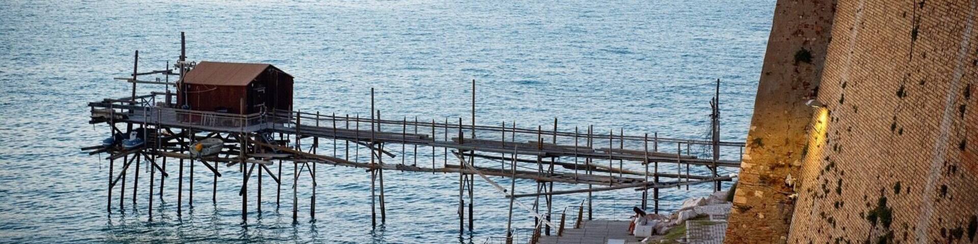 A pier extending into calm waters, a wooden structure on the pier, and a stone wall on the right.