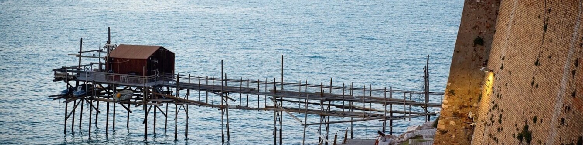 Un muelle que se adentra en aguas tranquilas, una estructura de madera sobre el muelle y un muro de piedra a la derecha.
