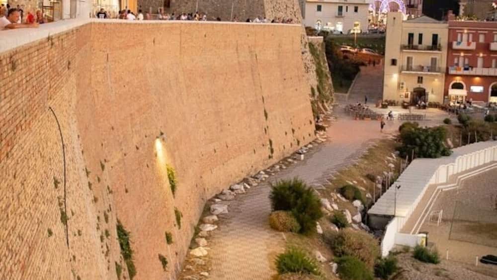 A historic stone wall with a tower, a paved pathway, and modern buildings in the background.