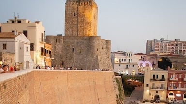 Un muro de piedra histórico con una torre, un camino empedrado y edificios modernos al fondo.