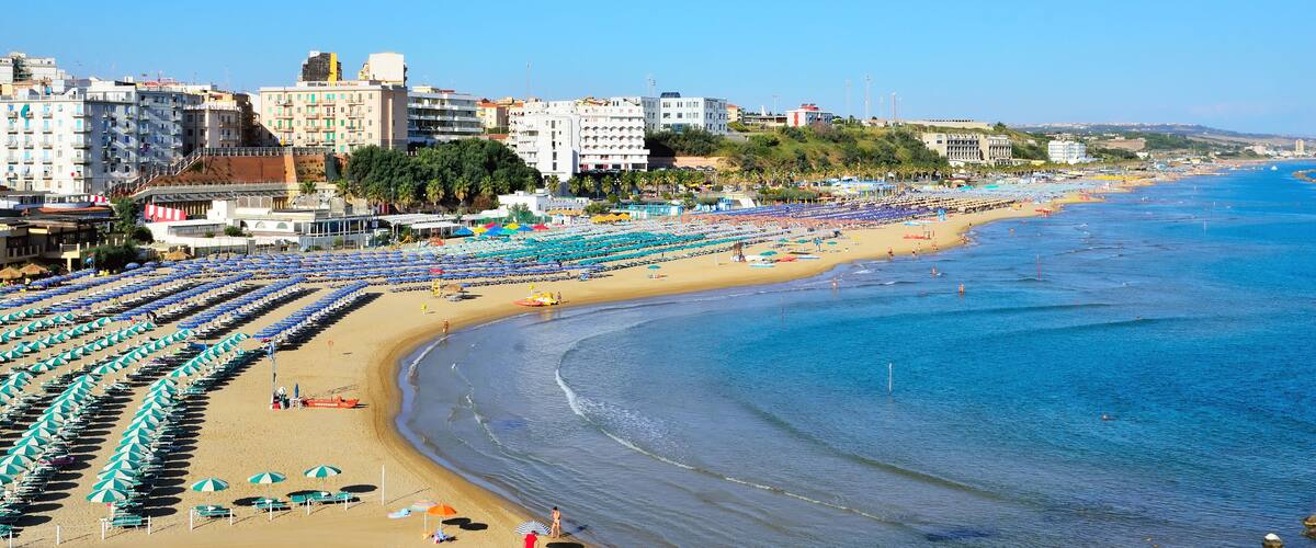A coastal cityscape with a wide beach lined with umbrellas and sun loungers, buildings, and a clear blue sea.