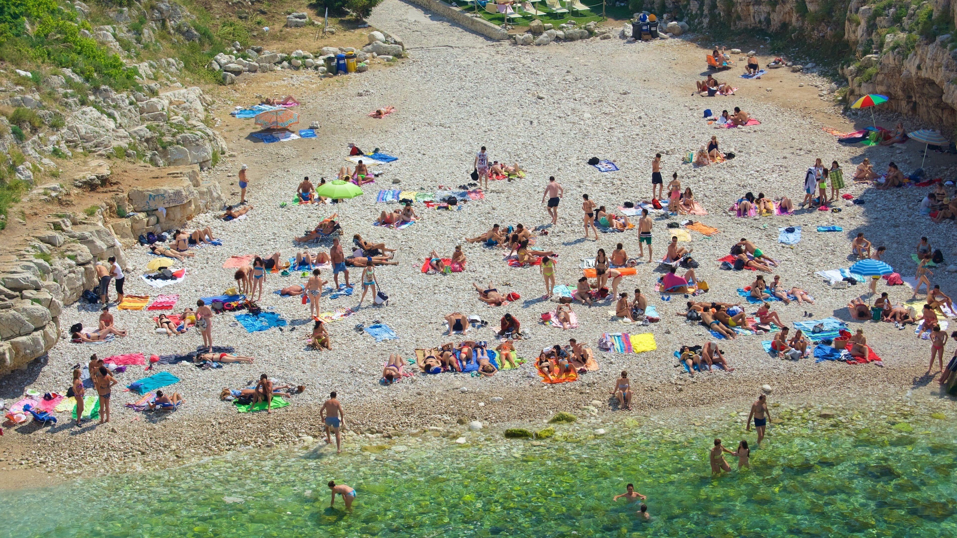 A crowded pebble beach with people sunbathing and swimming.