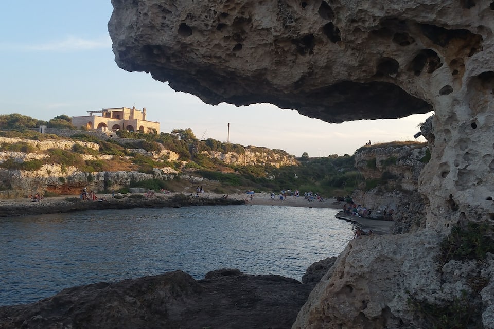 A coastal scene with a large rock formation, a beach, and a historic building on a hill.