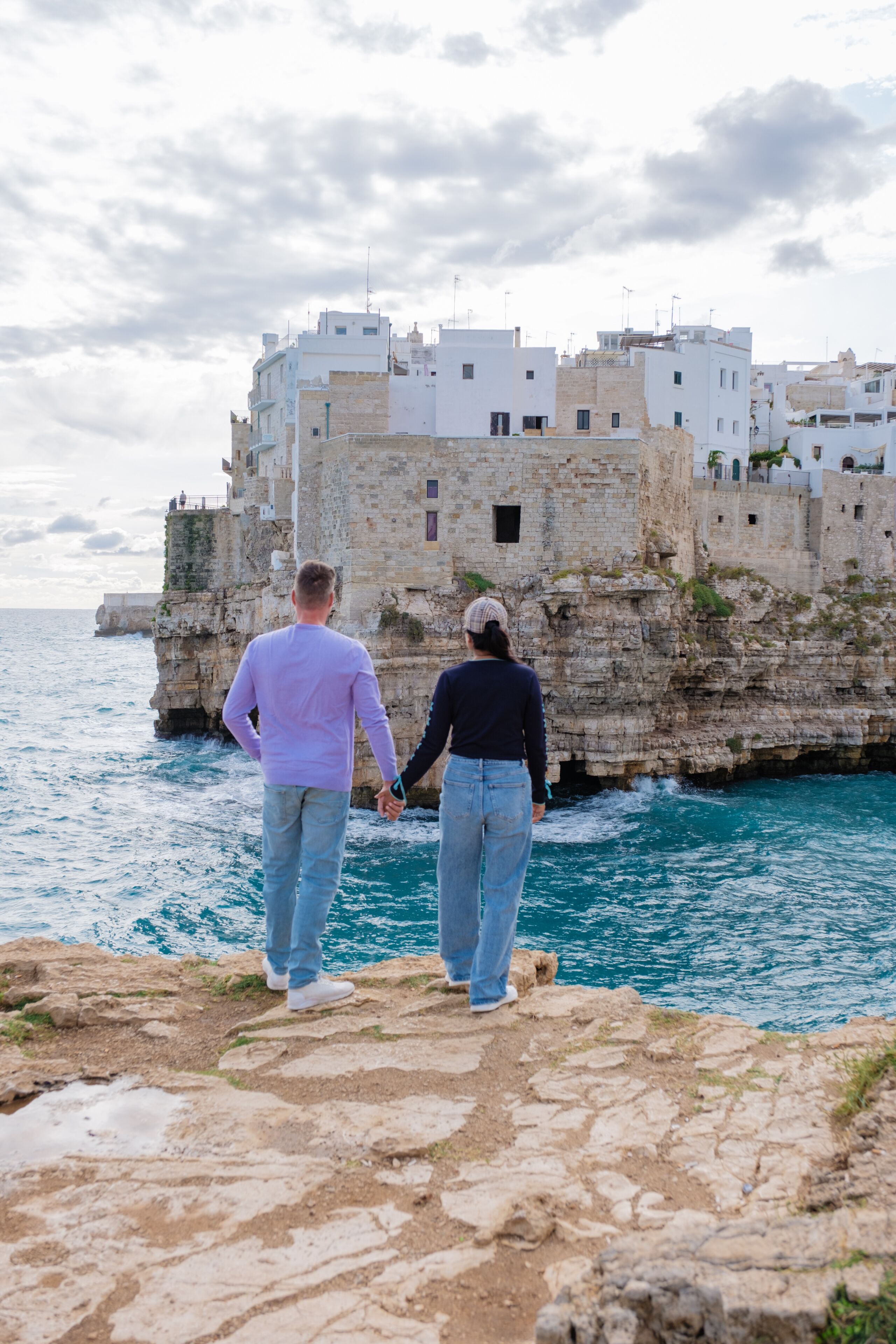 Una pareja caminando por un sendero rocoso frente al mar, con vistas a un edificio histórico de piedra con varios niveles y balcones.
