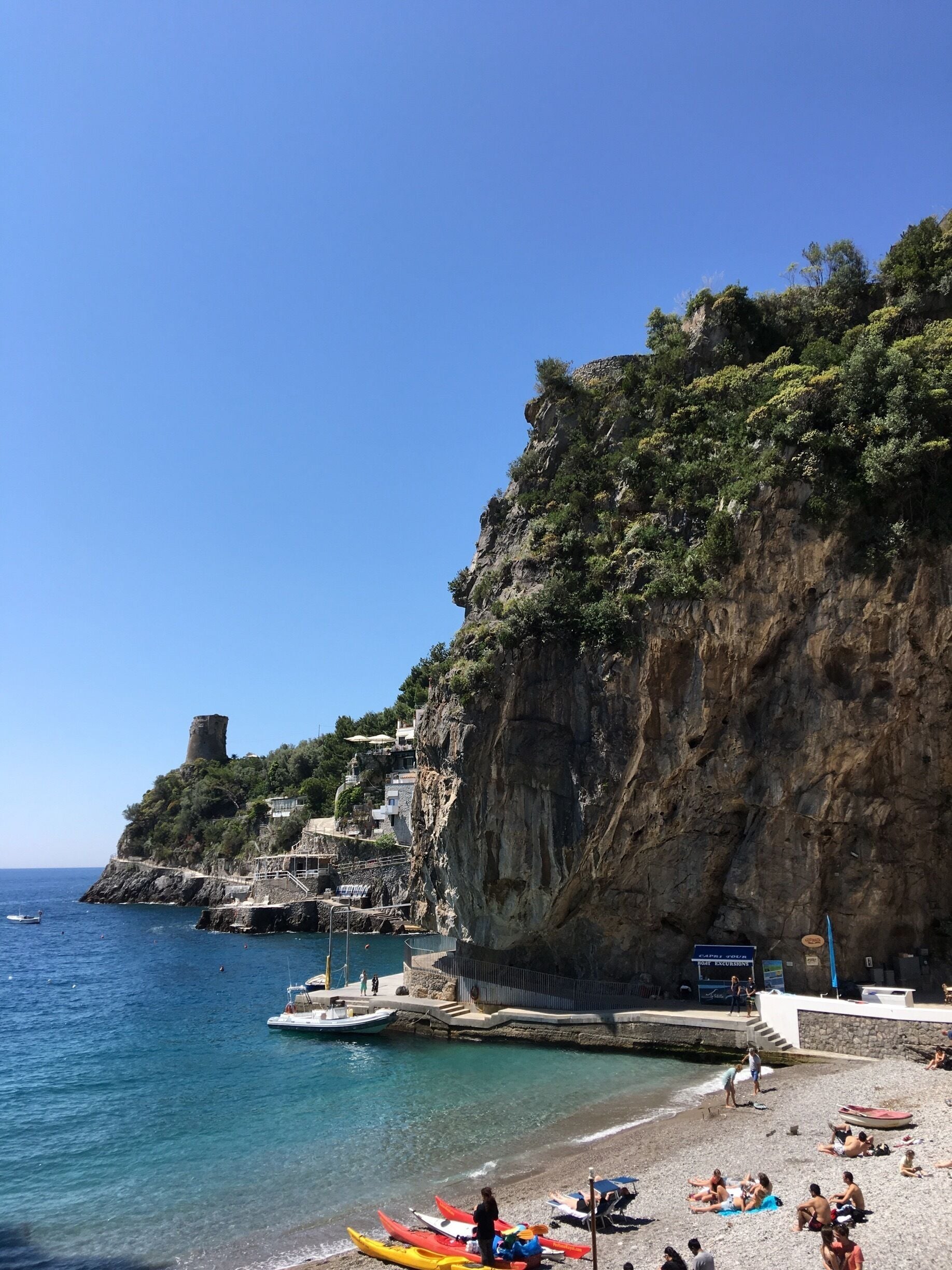 A coastal town with a historic tower, rocky cliffs, and a beach with people and boats.