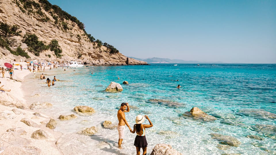 A beach with clear turquoise water, rocky shore, and a hilly backdrop.