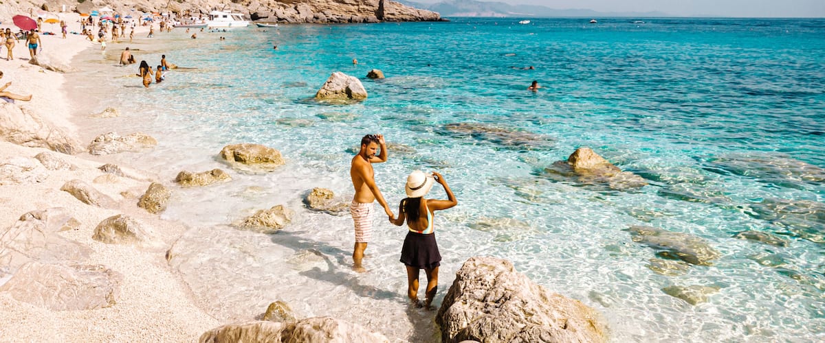 A beach with clear turquoise water, rocky shore, and a hilly backdrop.