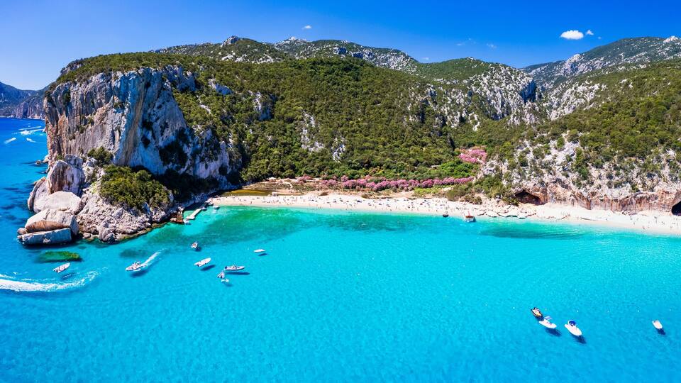 A coastal landscape with a beach, clear turquoise waters, boats, and rocky cliffs.