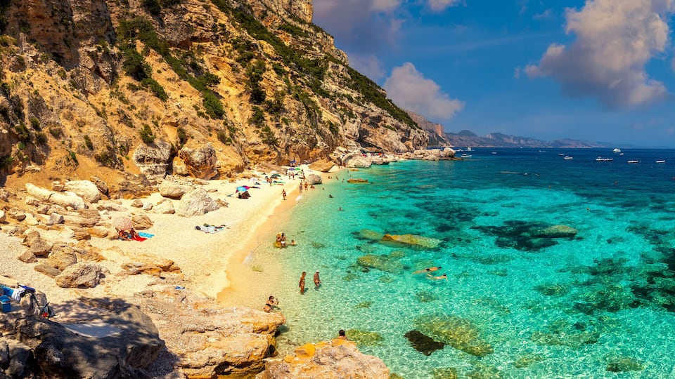 A rocky beach with clear turquoise water, a sandy shore, and boats in the distance.