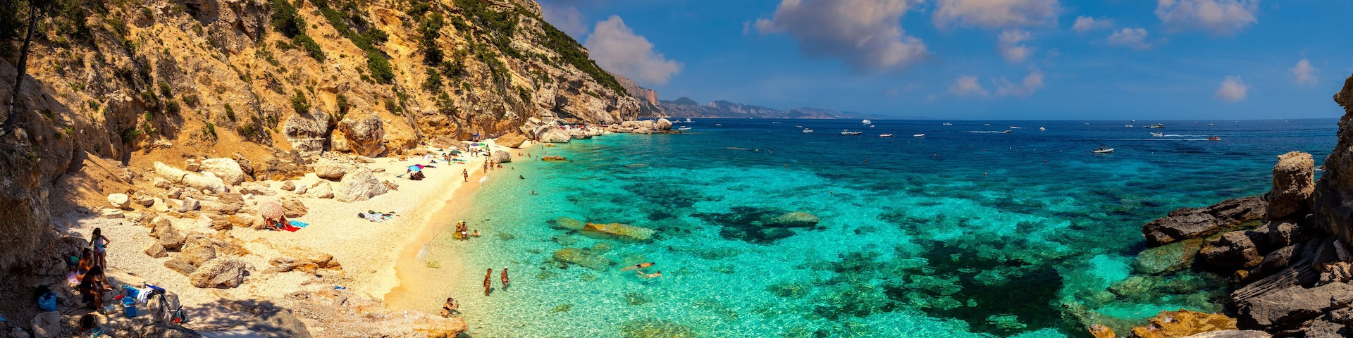 A rocky beach with clear turquoise water, a sandy shore, and boats in the distance.