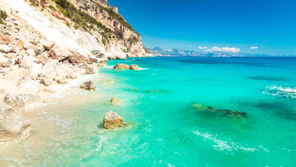 A coastal landscape with clear turquoise waters, rocky cliffs, and distant mountains.