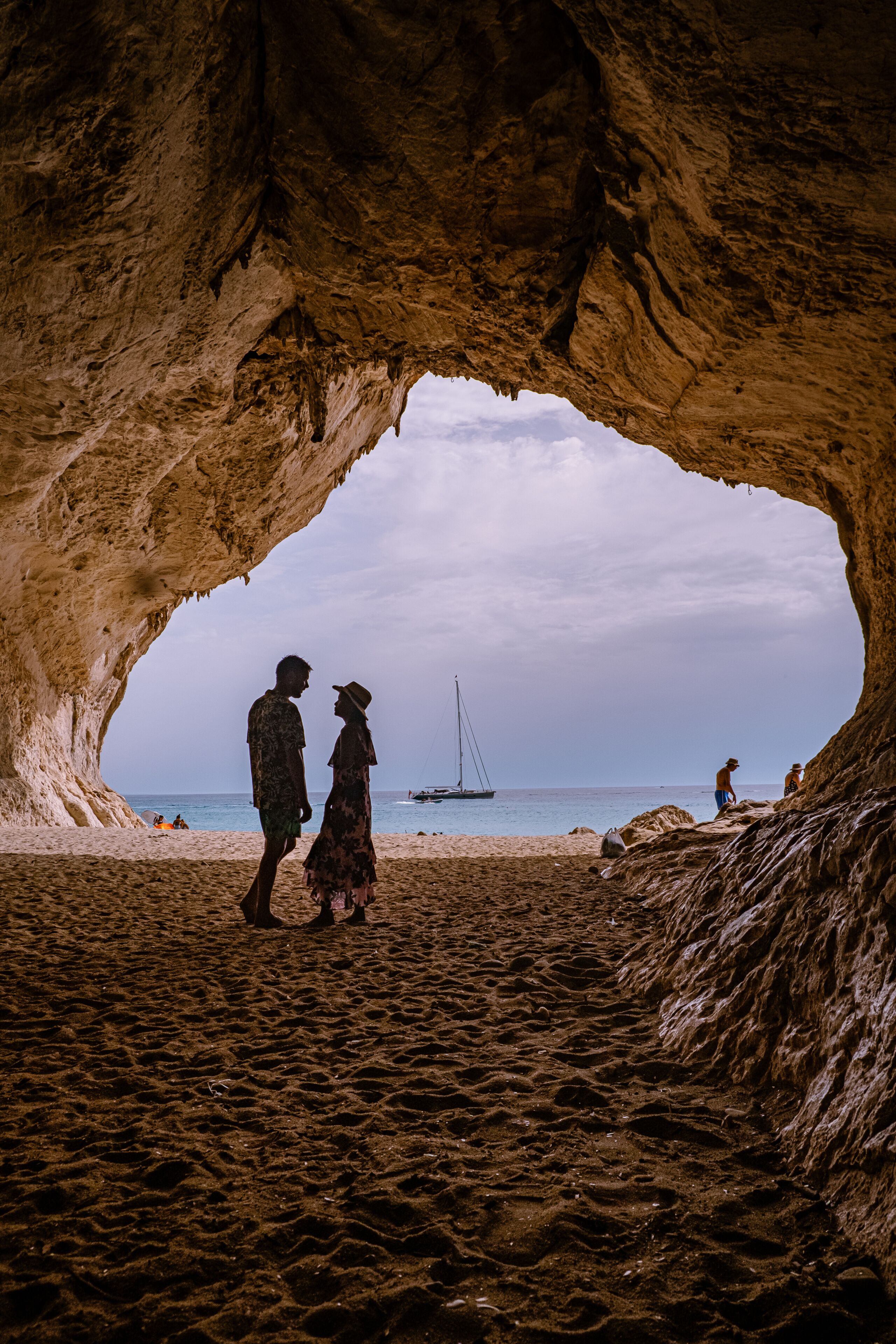 A cave opening to a beach with people walking and a sailboat in the distance.
