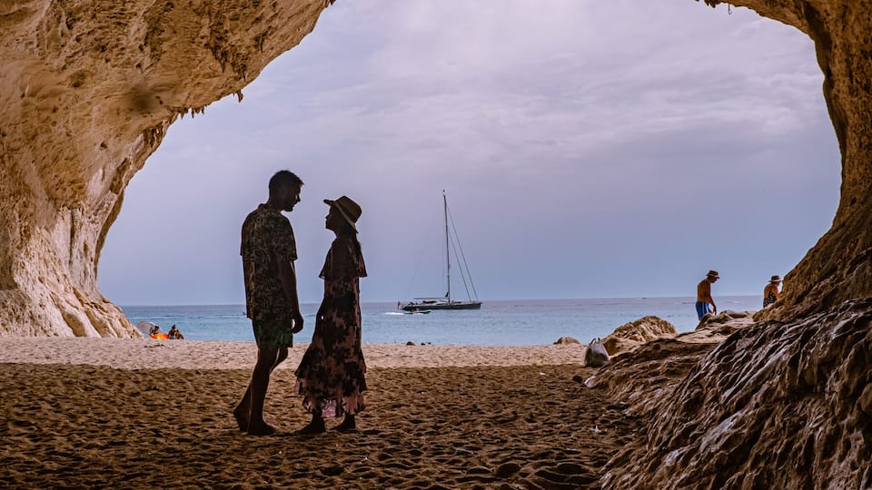 A cave opening to a beach with people walking and a sailboat in the distance.