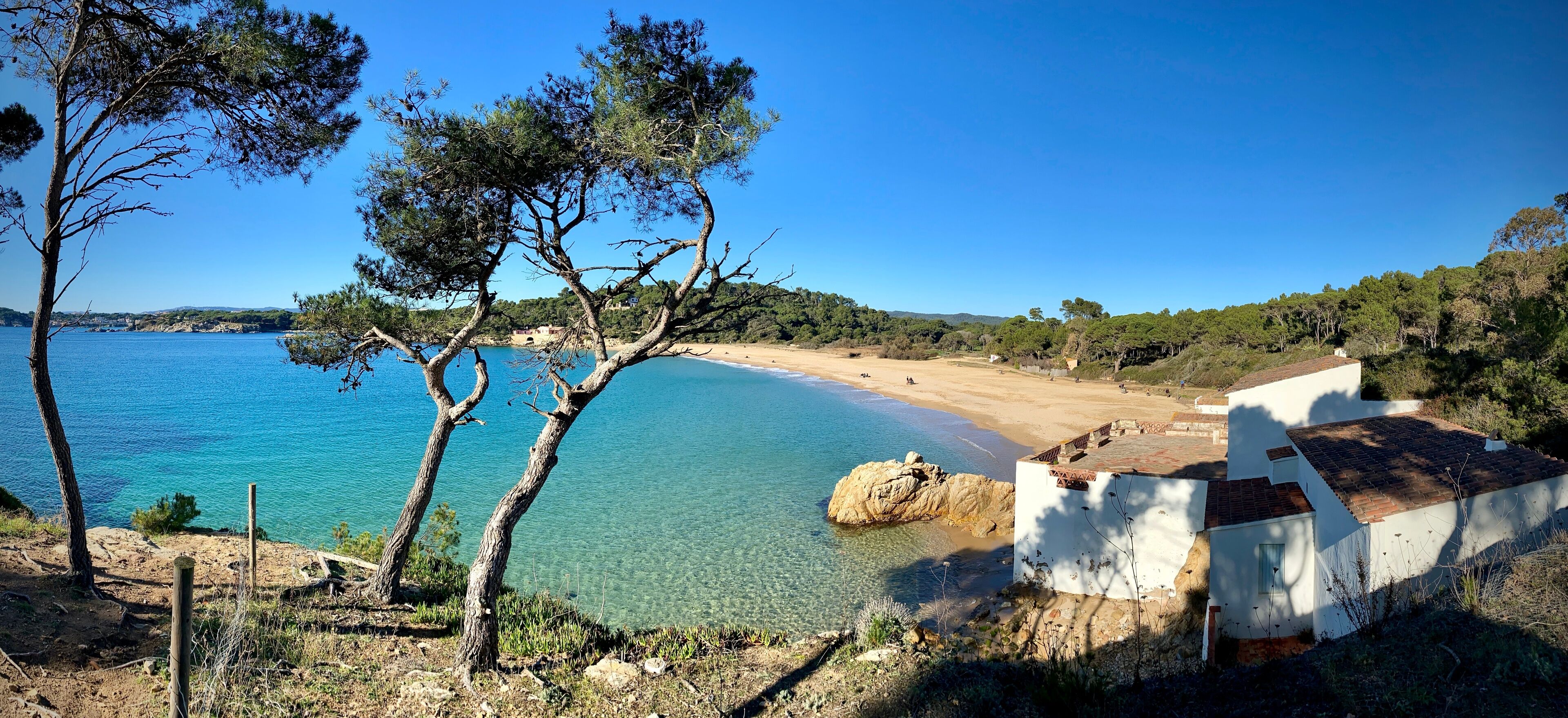 A coastal landscape with clear blue waters, a sandy beach, and a white building with a tiled roof.